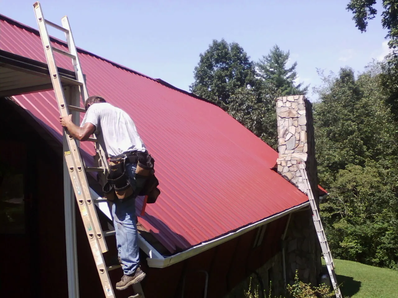 Roofer on a ladder beside a red metal roof and stone chimney — Soffit & Fascia Repair by Roofing Brothers in Adrian